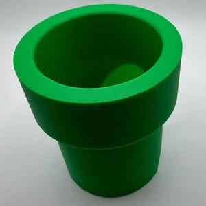 May include: A bright green, cylindrical pipe, reminiscent of a video game element. The pipe has a wide, rounded rim and a textured surface. The object is a solid, vibrant green color.