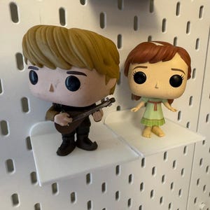 Pode incluir: Duas figuras de vinil pop em prateleiras brancas. A figura &agrave; esquerda &eacute; uma figura masculina com cabelo castanho tocando uma guitarra. A figura &agrave; direita &eacute; uma figura feminina com cabelo castanho vestindo um vestido verde.