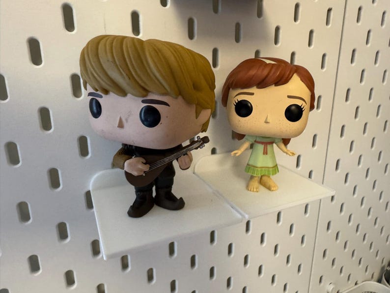 Pode incluir: Duas figuras de vinil pop em prateleiras brancas. A figura &agrave; esquerda &eacute; uma figura masculina com cabelo castanho tocando uma guitarra. A figura &agrave; direita &eacute; uma figura feminina com cabelo castanho vestindo um vestido verde.