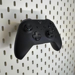 May include: A black Xbox controller with textured grips and several buttons. The controller includes a directional pad, two analogue sticks, and function buttons. It is set against a white pegboard background.