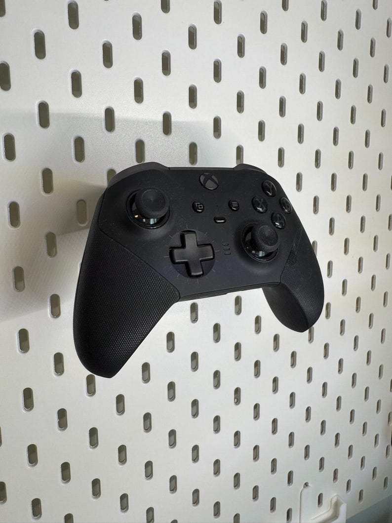May include: A black Xbox controller with textured grips and several buttons. The controller includes a directional pad, two analogue sticks, and function buttons. It is set against a white pegboard background.