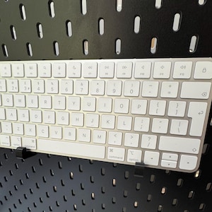 May include: A silver and white wireless keyboard with a full QWERTY layout. The keyboard is mounted on a black pegboard. The keys are white with black lettering and symbols. The keyboard has a sleek, modern design.