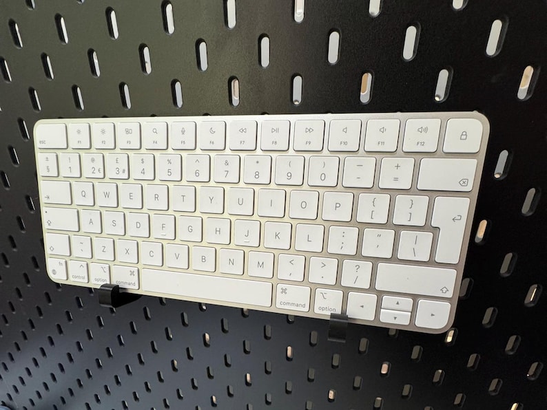 May include: A silver and white wireless keyboard with a full QWERTY layout. The keyboard is mounted on a black pegboard. The keys are white with black lettering and symbols. The keyboard has a sleek, modern design.