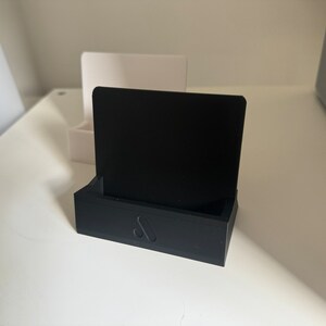 May include: A black 3D-printed phone stand with a rectangular base and a vertical back. The stand has a small logo on the front. A white stand is visible in the background.