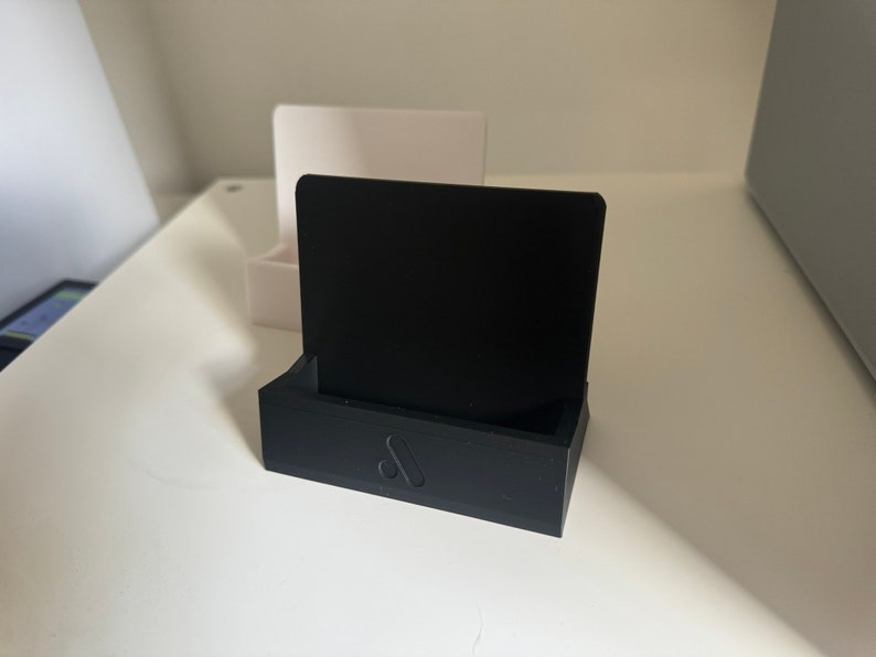 May include: A black 3D-printed phone stand with a rectangular base and a vertical back. The stand has a small logo on the front. A white stand is visible in the background.