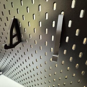 May include: Black pegboard with two black metal hooks. The pegboard has evenly spaced, oval-shaped holes. One hook is shaped like the letter "A", and the other is a straight bar with a bent end. The pegboard is mounted on a white surface.