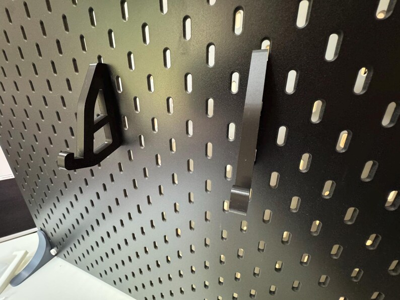 May include: Black pegboard with two black metal hooks. The pegboard has evenly spaced, oval-shaped holes. One hook is shaped like the letter "A", and the other is a straight bar with a bent end. The pegboard is mounted on a white surface.