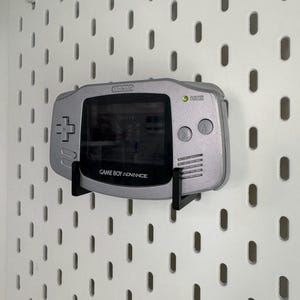 May include: A silver Nintendo Game Boy Advance handheld console mounted on a white pegboard. The screen displays a game. The console is held by a black bracket. The text "GAME BOY ADVANCE" is visible.