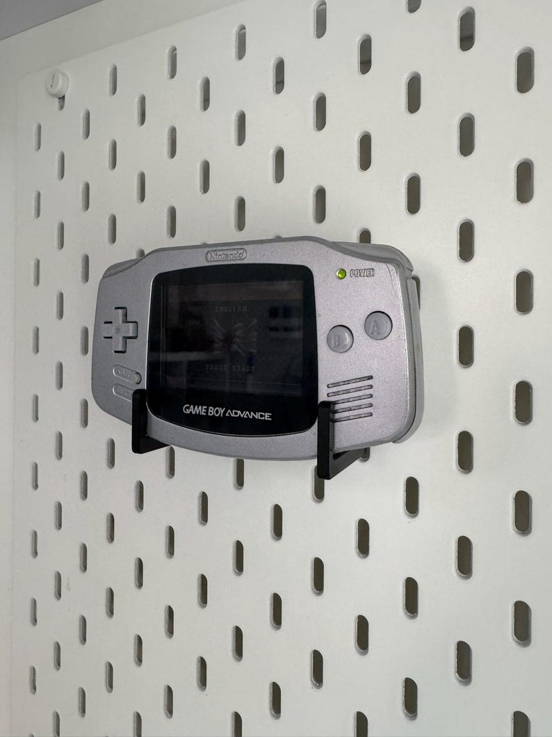 May include: A silver Nintendo Game Boy Advance handheld console mounted on a white pegboard. The screen displays a game. The console is held by a black bracket. The text "GAME BOY ADVANCE" is visible.