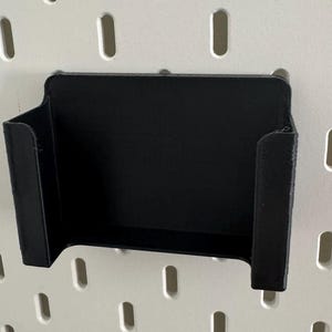 May include: A black plastic wall mount holder with a rectangular opening. The holder is designed to hold a small object, such as a phone or a tablet.
