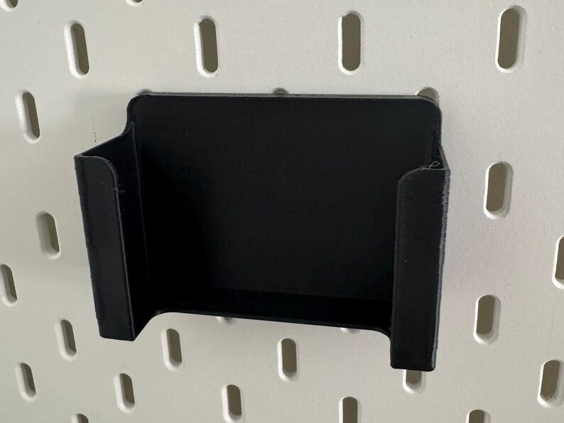 May include: A black plastic wall mount holder with a rectangular opening. The holder is designed to hold a small object, such as a phone or a tablet.