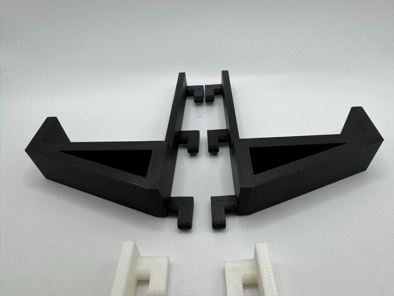 Op de afbeelding: Zwarte 3D-geprinte standaard met een driehoekige basis en een rechthoekige draagconstructie. De standaard is ontworpen om een ​​item vast te houden. Twee witte 3D-geprinte steunstukken zijn ook zichtbaar.