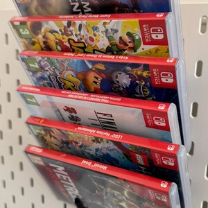 Pu&ograve; includere: Primo piano di uno scaffale a muro con sei cartucce di gioco per Nintendo Switch. Le cartucce sono disposte verticalmente, con la cartuccia superiore che &egrave; "Super Mario Party" e la cartuccia inferiore che &egrave; "Metroid Dread".