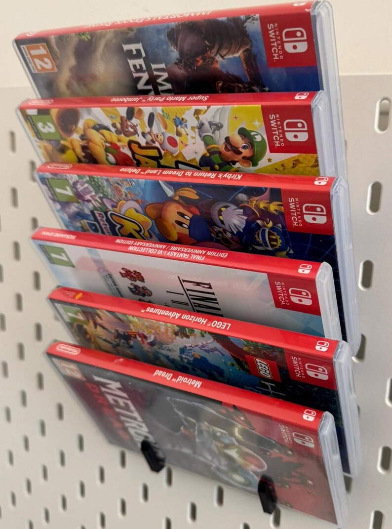 Pu&ograve; includere: Primo piano di uno scaffale a muro con sei cartucce di gioco per Nintendo Switch. Le cartucce sono disposte verticalmente, con la cartuccia superiore che &egrave; "Super Mario Party" e la cartuccia inferiore che &egrave; "Metroid Dread".