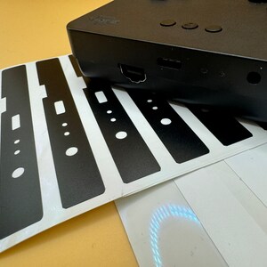 May include: A sheet of black vinyl stickers with cutouts for buttons and ports. The stickers are designed to cover the front of a device, such as a games console or media player.