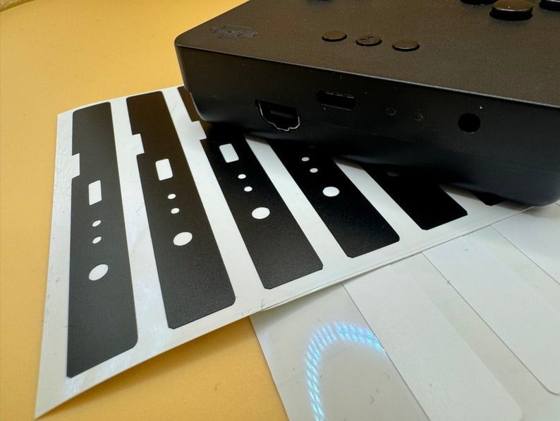 May include: A sheet of black vinyl stickers with cutouts for buttons and ports. The stickers are designed to cover the front of a device, such as a games console or media player.