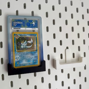 May include: A graded Pokémon card of Vaporeon, displayed in a protective case. The card features a blue and yellow design with the Pokémon character. The case is held in a black holder against a white pegboard.