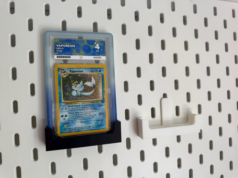 Op de afbeelding: Een beoordeelde Pok&eacute;mon-kaart van Vaporeon, tentoongesteld in een beschermende hoes. De kaart heeft een blauw en geel ontwerp met het Pok&eacute;mon-personage. De hoes wordt in een zwarte houder tegen een wit pegboard gehouden.
