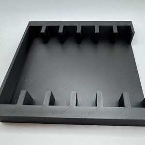 May include: A black, rectangular organizer with multiple vertical dividers. The organizer has a tray-like design with raised edges and is likely made of plastic or a similar material. The dividers create slots for storing items.