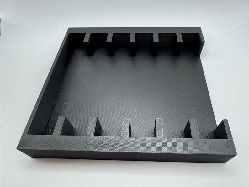 May include: A black, rectangular organizer with multiple vertical dividers. The organizer has a tray-like design with raised edges and is likely made of plastic or a similar material. The dividers create slots for storing items.