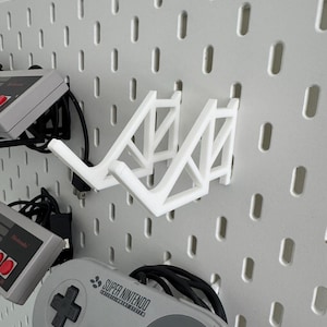 Puede incluir: Soporte de pared blanco impreso en 3D para dos mandos de videojuegos. El soporte est&aacute; fijado a una pared de tablero de clavijas blanco. Un mando Super Nintendo gris est&aacute; colgado del soporte. El mando tiene el texto "SUPER NINTENDO" impreso en &eacute;l.