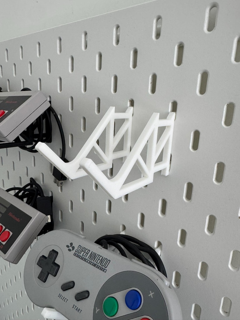 Puede incluir: Soporte de pared blanco impreso en 3D para dos mandos de videojuegos. El soporte est&aacute; fijado a una pared de tablero de clavijas blanco. Un mando Super Nintendo gris est&aacute; colgado del soporte. El mando tiene el texto "SUPER NINTENDO" impreso en &eacute;l.