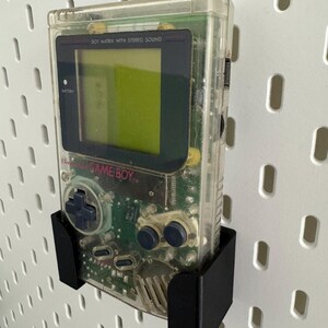 Op de afbeelding: Een transparante Game Boy Color handheld-videogamesysteem met een zwarte wandbeugel. De console heeft een groen scherm en de tekst "DOT MATRIX WITH STEREO SOUND" en "GAME BOY" is zichtbaar.