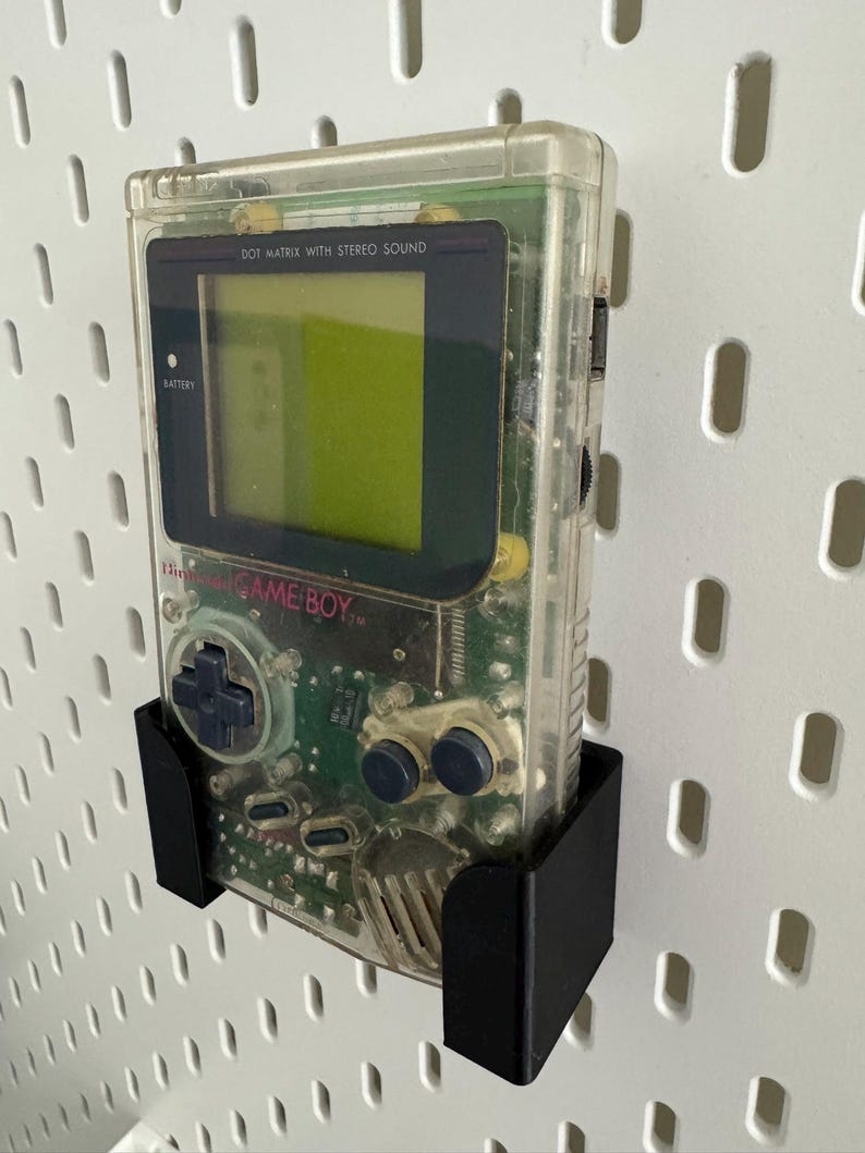 Op de afbeelding: Een transparante Game Boy Color handheld-videogamesysteem met een zwarte wandbeugel. De console heeft een groen scherm en de tekst "DOT MATRIX WITH STEREO SOUND" en "GAME BOY" is zichtbaar.