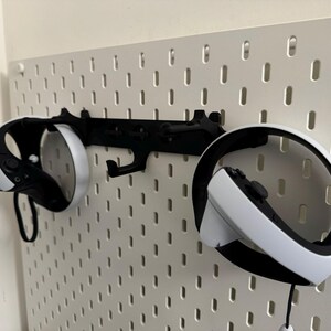 May include: Black VR headset and controllers mounted on a white pegboard. The controllers are white and black, and the headset is black with white accents. The pegboard is mounted on a white wall.