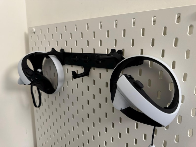May include: Black VR headset and controllers mounted on a white pegboard. The controllers are white and black, and the headset is black with white accents. The pegboard is mounted on a white wall.