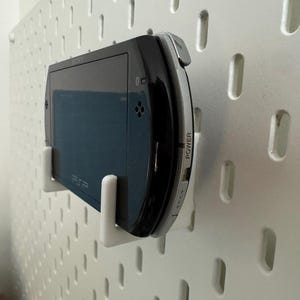 May include: A black and silver handheld gaming console, mounted on a white pegboard with white brackets. The console has a screen and buttons, with the word "POWER" visible on the side. The console is a PSP.