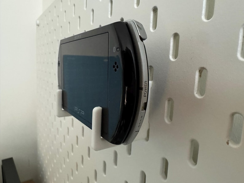 May include: A black and silver handheld gaming console, mounted on a white pegboard with white brackets. The console has a screen and buttons, with the word "POWER" visible on the side. The console is a PSP.