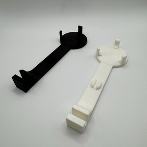 K&ouml;nnte beinhalten: Zwei 3D-gedruckte Handyhalter, einer schwarz und einer wei&szlig;. Beide Halter haben eine runde Basis mit drei Zacken und einen langen Griff mit einem Haken am Ende.