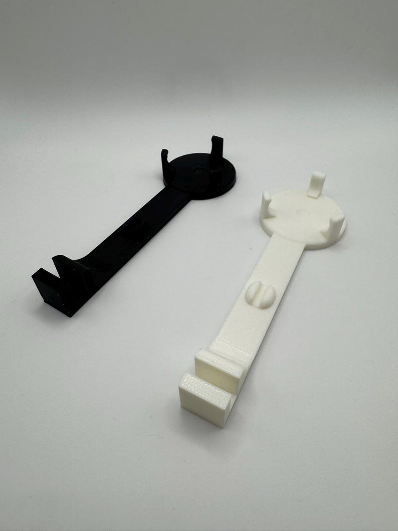 K&ouml;nnte beinhalten: Zwei 3D-gedruckte Handyhalter, einer schwarz und einer wei&szlig;. Beide Halter haben eine runde Basis mit drei Zacken und einen langen Griff mit einem Haken am Ende.