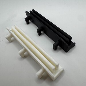 Pode incluir: Dois objetos de pl&aacute;stico impressos em 3D. Um &eacute; branco, o outro preto. Ambos t&ecirc;m uma base retangular com duas barras paralelas elevadas e tr&ecirc;s suportes perpendiculares. O objeto branco &eacute; ligeiramente esbranqui&ccedil;ado, enquanto o preto &eacute; preto s&oacute;lido.