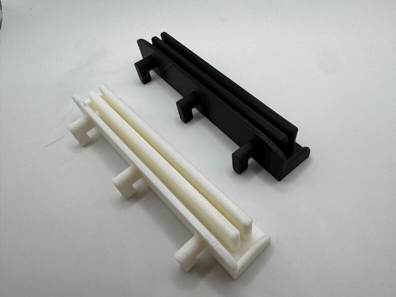 Pode incluir: Dois objetos de pl&aacute;stico impressos em 3D. Um &eacute; branco, o outro preto. Ambos t&ecirc;m uma base retangular com duas barras paralelas elevadas e tr&ecirc;s suportes perpendiculares. O objeto branco &eacute; ligeiramente esbranqui&ccedil;ado, enquanto o preto &eacute; preto s&oacute;lido.