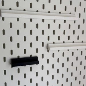 以下が含まれることがあります： 白のペグボードに3つの棚：白2つと黒1つ。棚は長方形で、ペグボードの楕円形の穴に合うように設計されています。ペグボードは白で、楕円形の穴が均等に配置されています。