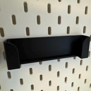 Puede incluir: Un estante negro rectangular con lados curvos est&aacute; montado en una pared perforada blanca. El estante est&aacute; dise&ntilde;ado para guardar objetos peque&ntilde;os. La pared perforada tiene agujeros ovalados espaciados uniformemente.