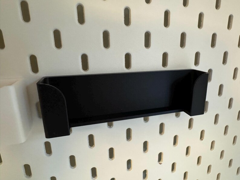 Puede incluir: Un estante negro rectangular con lados curvos est&aacute; montado en una pared perforada blanca. El estante est&aacute; dise&ntilde;ado para guardar objetos peque&ntilde;os. La pared perforada tiene agujeros ovalados espaciados uniformemente.