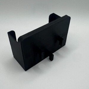 May include: A black plastic 3D printed phone stand with two slots for holding a phone. The stand has a rectangular base and a raised back.