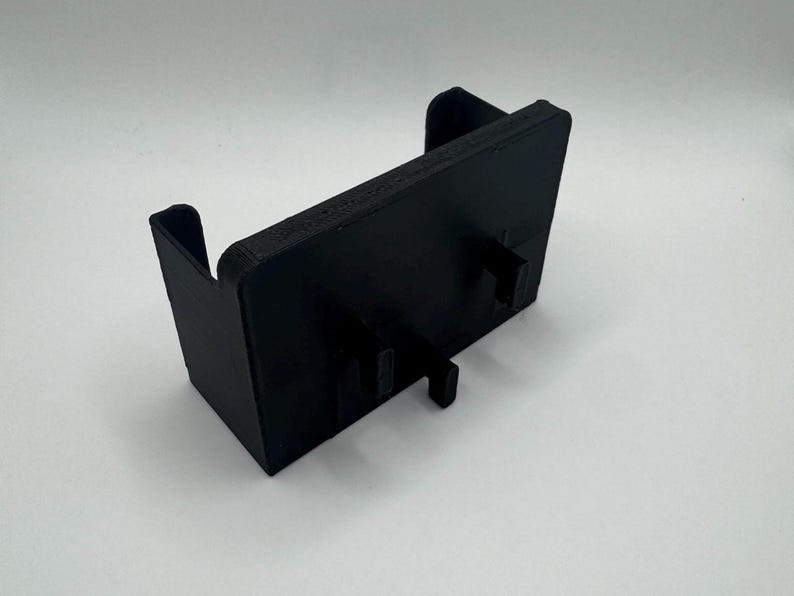 May include: A black plastic 3D printed phone stand with two slots for holding a phone. The stand has a rectangular base and a raised back.