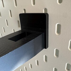 K&ouml;nnte beinhalten: Ein schwarzer Regalhalter f&uuml;r Lochw&auml;nde, zur Befestigung an einer Lochwand. Der Halter besteht aus einem soliden schwarzen Material und verf&uuml;gt &uuml;ber eine rechteckige Regalst&uuml;tze. Er ist an einer wei&szlig;en Lochwand mit ovalen L&ouml;chern befestigt.