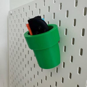 May include: A bright green, 3D-printed pipe-shaped organizer, reminiscent of a video game, is mounted on a white pegboard. The pipe holds various small items, including black and orange objects.
