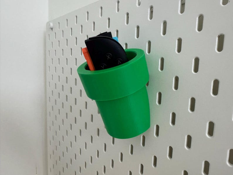 May include: A bright green, 3D-printed pipe-shaped organizer, reminiscent of a video game, is mounted on a white pegboard. The pipe holds various small items, including black and orange objects.
