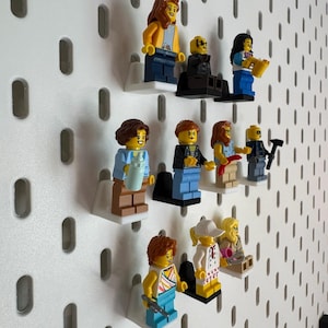 K&ouml;nnte beinhalten: Eine Sammlung von Lego-Minifiguren, die auf einer wei&szlig;en Lochwand ausgestellt sind. Die Minifiguren tragen verschiedene Outfits und Accessoires, darunter Werkzeuge, Getr&auml;nke und Musikinstrumente. Die Figuren sind auf kleinen wei&szlig;en Regalen montiert.