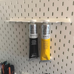 May include: Two tubes of Winsor & Newton Winton Oil Colour paint, one black and one yellow, displayed on a white pegboard shelf. The tubes are silver with black and yellow labels. The pegboard has a grid of evenly spaced holes.