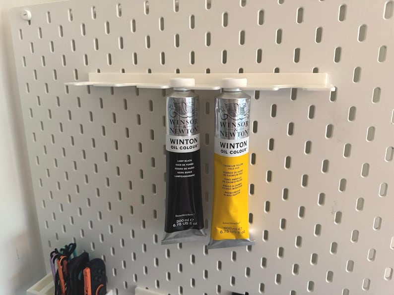 May include: Two tubes of Winsor & Newton Winton Oil Colour paint, one black and one yellow, displayed on a white pegboard shelf. The tubes are silver with black and yellow labels. The pegboard has a grid of evenly spaced holes.