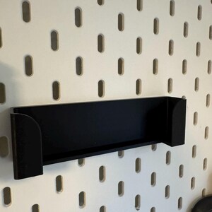 Puede incluir: Un estante negro con lados curvos est&aacute; montado en una pared perforada blanca. El estante es rectangular con una base plana y est&aacute; dise&ntilde;ado para sostener objetos. La pared perforada tiene agujeros ovalados espaciados.