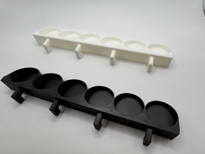 以下が含まれることがあります： 白と黒の2つの3Dプリントトレイ。それぞれに5つの円形のくぼみと、5つの小さな下向きのフックが付いています。これらのトレイは、小さなアイテムを保持するように設計されています。