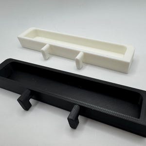 May include: Two rectangular, open-top trays with integrated hooks. One tray is black, the other is white. Both have a similar design, with a long, narrow shape and two protruding hooks on the underside for hanging.